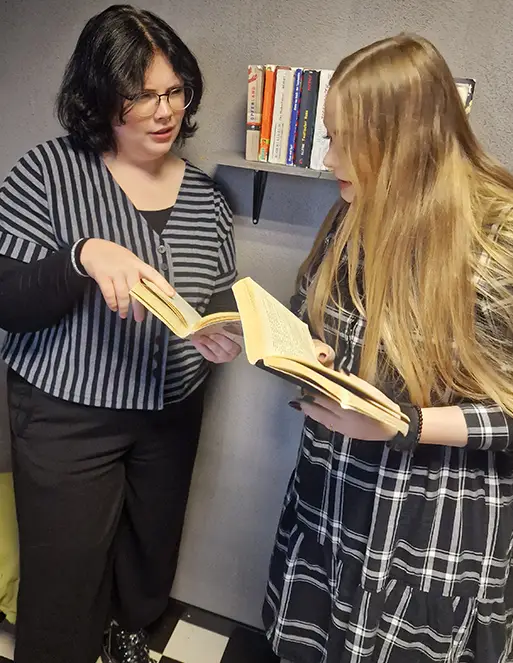 Auf dem Bild sind zwei Frau. Die blonde Frau hat ein Buch in der Hand und schaut in das Buch von der Frau mit der Brille. Die Frau mit der Brille zeigt dabei in das Buch und schaut die blonde Frau an.