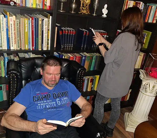 Ein Mann sitzt in einem schwarzen Sessel und ließt ein Buch. Hinter ihm ist ein Bücherregal an dem eine Frau steht und sich die Bücher anschaut.