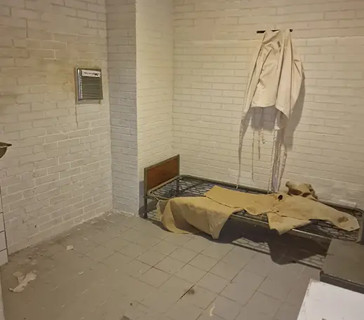 In dem weißen Raum steht ein Bett ohne Matratze. Auf dem Bett liegt eine ausgebreitete Jacke. Über dem Bett hängt ein weißer Kittel an einem Haken.